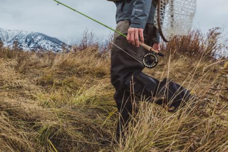 A fisherman carries a fishing rod through tall grass near mountains in Jackson Hole