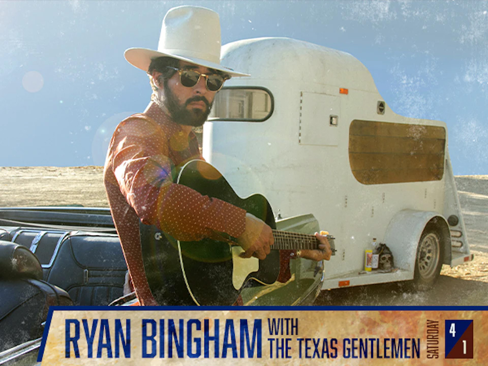 Ryan Bingham playing an acoustic guitar while leaning against a convertible car towing a white trailer
