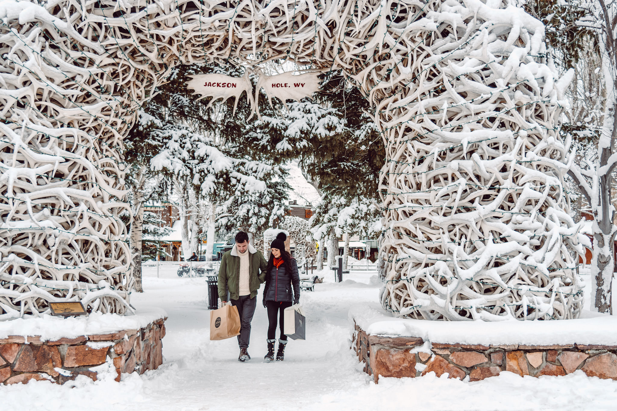 A couple bundled up in winter jackets and boots walk under the snowy antler arch at Town Square