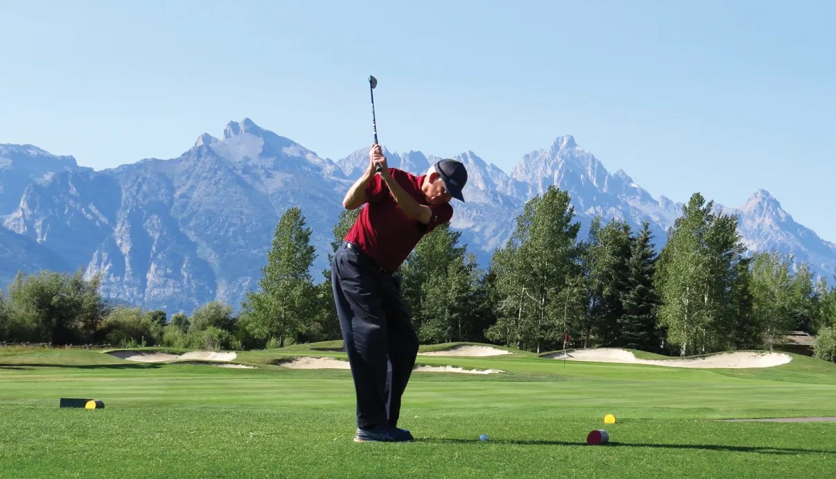 a man swings a golf club on a pristine golf course with the Tetons in the distance